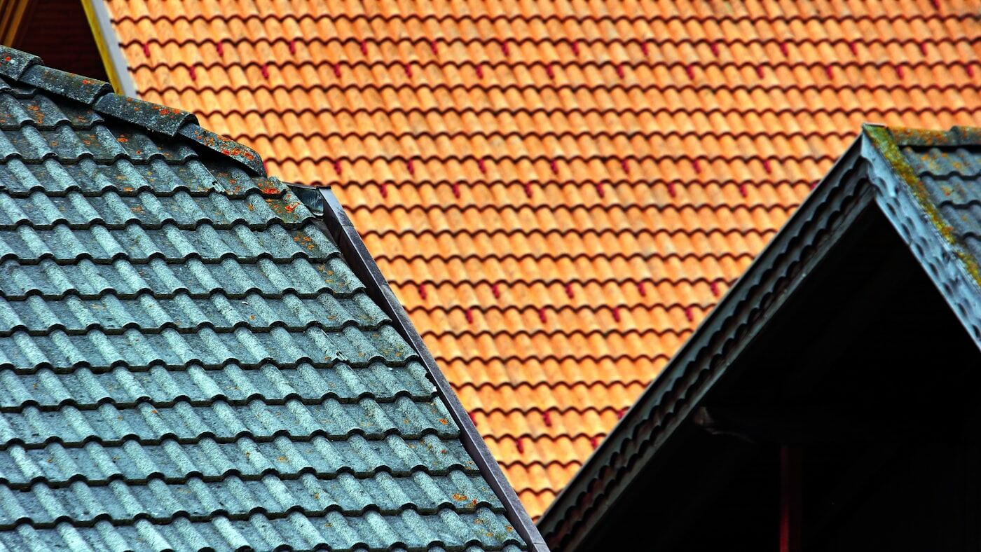 Twee daken die tegen elkaar aan liggen: het ene met oranje keramische pannen, het andere met blauwgrijze pannen